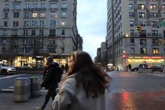 two people walking down the street in front of some tall buildings at dusk with lights on