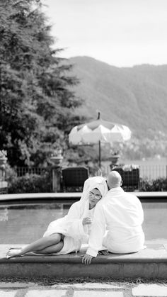 a man and woman sitting on the ground next to a pool with umbrellas in the background