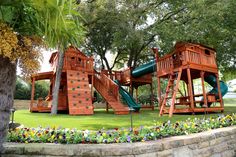 a wooden play set in the middle of a park with trees and flowers around it