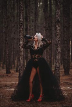 a woman with white hair and black dress standing in the woods holding a wine bottle