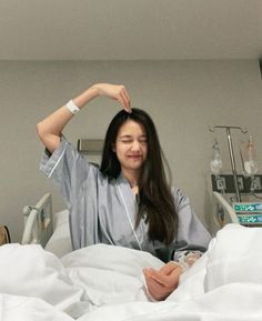 a woman sitting in a hospital bed holding her hair up to her head and smiling