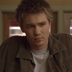 a young man with blonde hair wearing a brown jacket and white t - shirt looking at the camera