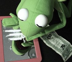 a green stuffed animal sitting on top of a red tablet next to a pile of money