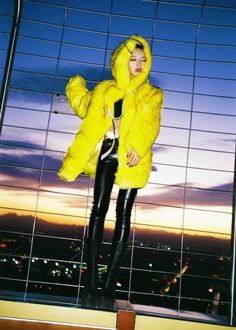 a woman standing in front of a window wearing a yellow fur coat and black leather pants