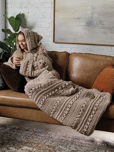 a woman sitting on top of a couch covered in a knitted blanket and holding a cup