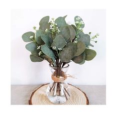 a glass vase filled with green leaves and white flowers on top of a wooden slice