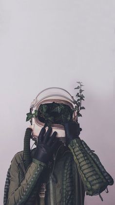 a person wearing black gloves and green jacket covering their face with plants growing out of it