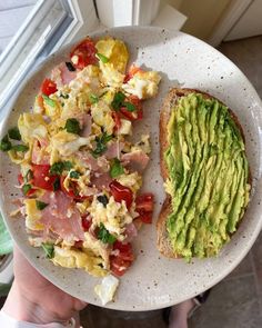 someone holding a plate with an avocado and scrambled eggs on it next to a slice of bread