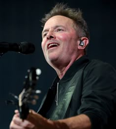 a man singing into a microphone while holding a guitar in his right hand and wearing earbuds