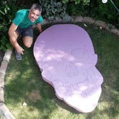 a man kneeling down next to a large purple skull shaped table in the grass with writing on it