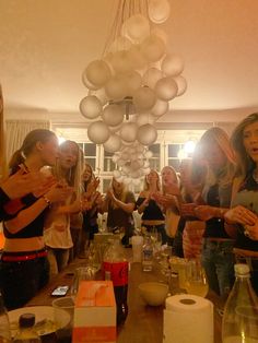 a group of women standing around a table with food and drinks on it, in front of a chandelier hanging from the ceiling