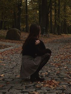 a woman kneeling down in the middle of a park with leaves on the ground and trees behind her