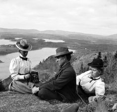 two men and a woman sitting on top of a hill next to each other near a lake