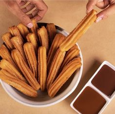 a bowl filled with churros next to dipping sauce and two hands reaching for one