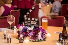 Movie clacker surrounded by purple floral as a reception table centerpiece for a movie wedding theme Movie Wedding Theme, Cinema Ideas, Loews Royal Pacific Resort, Ballroom Reception