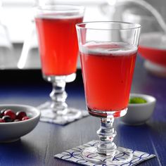 two glasses filled with red liquid sitting on top of a table next to bowls of cherries