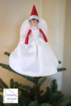 an elf doll sitting on top of a christmas tree in a white dress and red hat