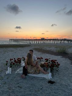 a man and woman sitting on the beach at sunset with flowers in front of them