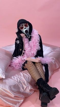 a person in a mask sitting on a bed with pink feathers and black stockings, holding a cup