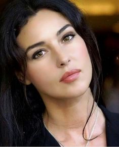 a woman with long black hair wearing a necklace and looking at the camera while standing in front of a store window