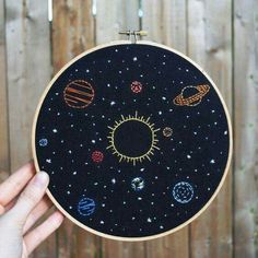 a person holding up an embroidery project with planets and sun in the center, on top of a wooden fence