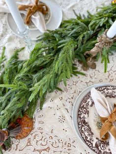 the table is set for christmas dinner with white plates and napkins, silverware and evergreen branches