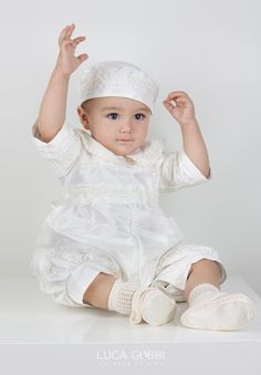 a baby sitting on the floor wearing a white dress and hat with one hand up in the air