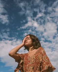 a woman standing under a cloudy blue sky with her hands on her head looking up