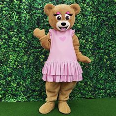 a brown teddy bear dressed in pink dress standing next to a green wall with bushes behind it