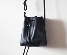 "This tiny bucket bag is designed to carry just a few items such as a small wallet, phone and keys. It features soft black vegan (fake) leather, an adjustable shoulder strap and a drawstring closure. Please note that this bag is very small. It will not fit large wallets. Details: -Dimensions: 7\" length (top to bottom), 10.5\" top width and 6\" bottom width (side to side), 4.5\" depth, 6\" X 4.5\" base (bottom) -Shoulder strap measures approx. 45-50\" total (adjustable) -Soft black faux (fake) l Minimalist Black Bucket Bag With Detachable Strap, Chic Bucket Bag With Adjustable Strap For Gift, Chic Bucket Bag With Adjustable Strap As A Gift, Black Pouch Bucket Bag As Gift, Black Shoulder Bucket Bag Gift, Black Bucket Bag With Adjustable Strap As Gift, Black Bucket Bag With Adjustable Strap For Gift, Large Wallet, Black Leather Crossbody Bag