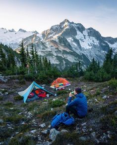 a man sitting on top of a mountain next to a tent and camping gear in the wilderness