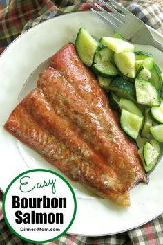 a white plate topped with salmon and cucumbers next to a bun on a table