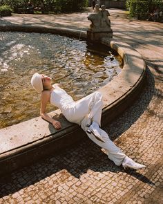 a woman laying on the ground in front of a fountain wearing a white suit and hat