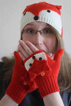 a woman wearing red gloves and a knit hat covers her face with both hands while looking at the camera