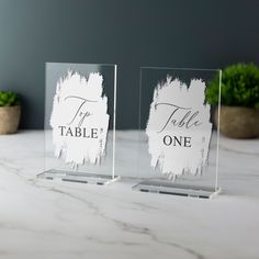 two clear acrylic signs with the words table one on them sitting on a marble surface