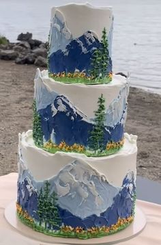 a three tiered cake decorated with mountains and pine trees on a table next to the water