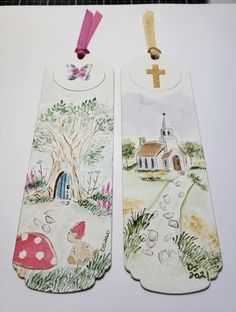 two bookmarks with designs on them are hanging from a cord and one has a small house in the background