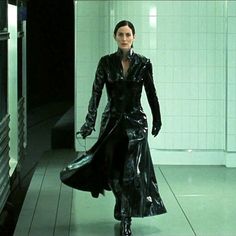 a woman in a black trench coat walking down a hallway