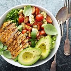 a white bowl filled with chicken, avocado, tomatoes and sliced cucumbers