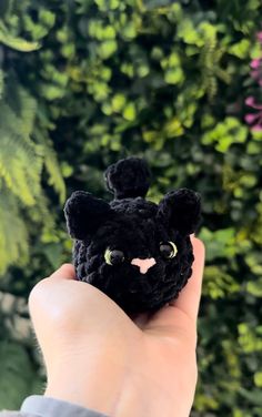 a hand holding a small black crocheted animal in front of a green hedge