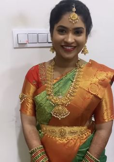 a woman in an orange and green sari with gold jewelry on her neck, standing next to a white wall