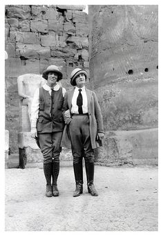 an old black and white photo of two people standing next to each other in front of a stone building