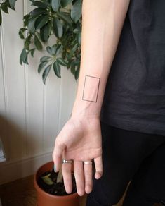 a person's hand with a small square tattoo on their left wrist, next to a potted plant