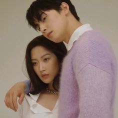 two people standing next to each other in front of a white wall and one person wearing a purple sweater