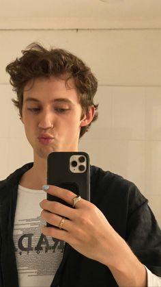 a young man looking at his cell phone in the bathroom while wearing a ring and black cardigan