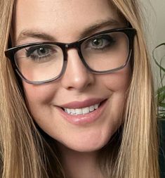 a close up of a person wearing glasses and smiling at the camera with a plant in the background