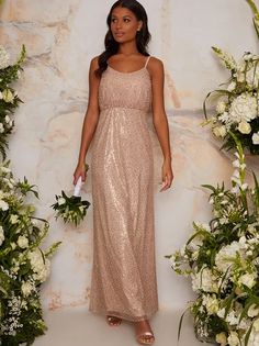 a woman standing in front of flowers wearing a gold sequin gown with spaghetti straps