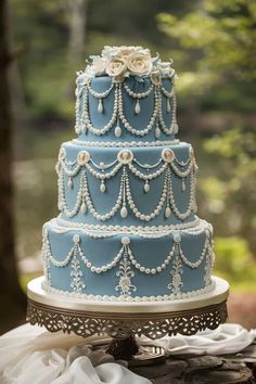 a three tiered blue wedding cake with pearls and flowers on the top is sitting on a table