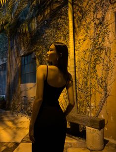 a woman in a black dress is standing near a bench and palm trees at night