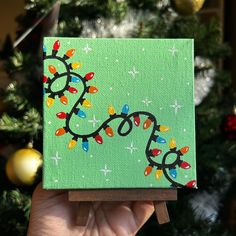 a hand holding up a small green box with christmas lights on it and a tree in the background